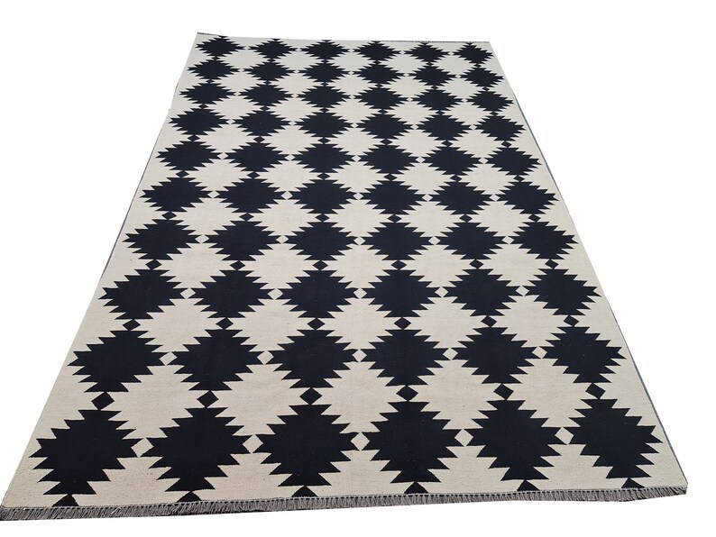 Puede incluir: Una alfombra rectangular con un patr&oacute;n repetido de formas de diamante negras sobre un fondo crema. La alfombra tiene un dise&ntilde;o geom&eacute;trico simple, adecuado para varios estilos de interior. La alfombra est&aacute; hecha de un material tejido.