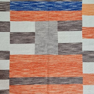 May include: A woven rug with a geometric pattern. The design features alternating blocks of color, including orange, blue, gray, and white. The rug has a textured appearance, with visible woven threads. The overall aesthetic is modern and abstract.