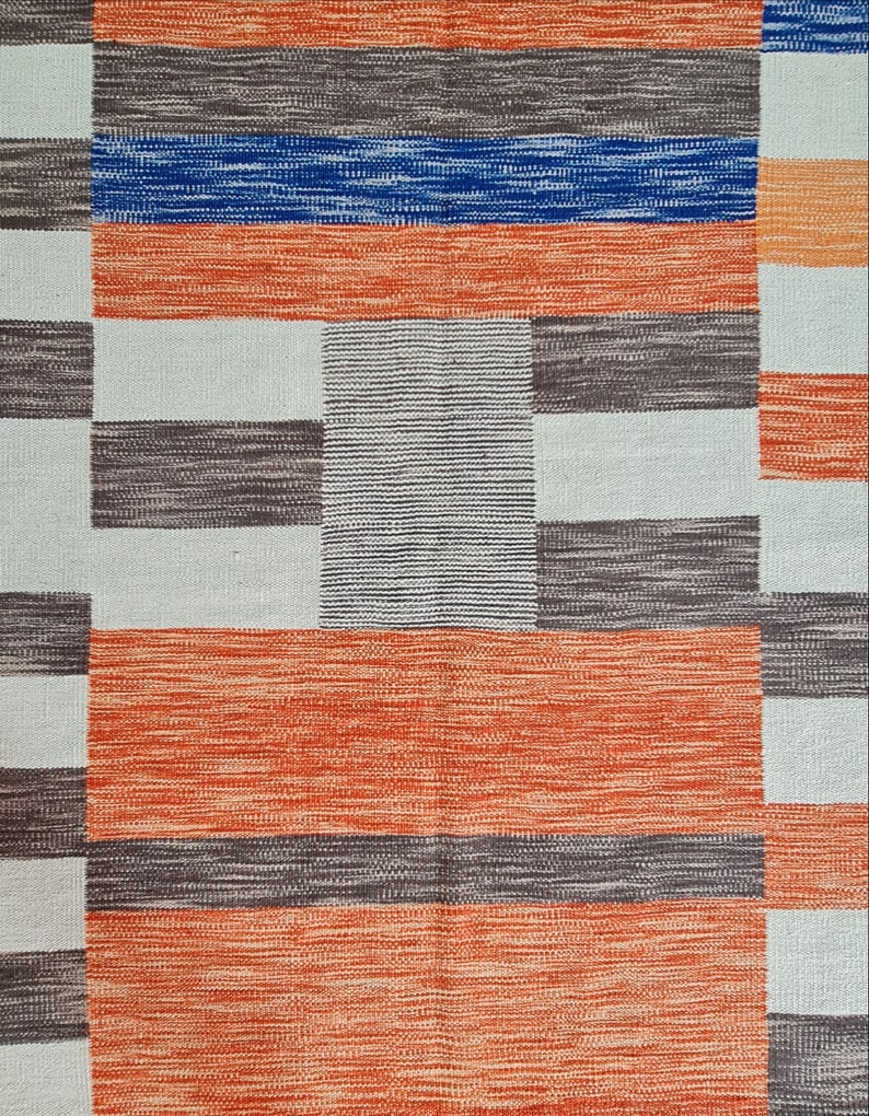 May include: A woven rug with a geometric pattern. The design features alternating blocks of color, including orange, blue, gray, and white. The rug has a textured appearance, with visible woven threads. The overall aesthetic is modern and abstract.
