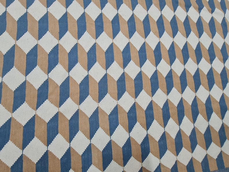 May include: A geometric patterned rug featuring a repeating 3D cube design. The rug's pattern uses white, blue, and tan colors. The rug is made of a woven material and is suitable for home decor.