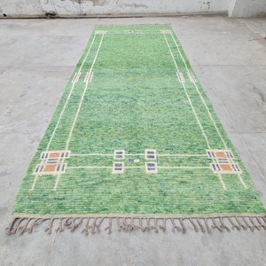 Puede incluir: Una alfombra rectangular verde con un dise&ntilde;o geom&eacute;trico. La alfombra presenta un campo verde claro con detalles en blanco, naranja y azul marino. La alfombra tiene flecos en los extremos cortos. La alfombra mide unos 152 cm de largo.