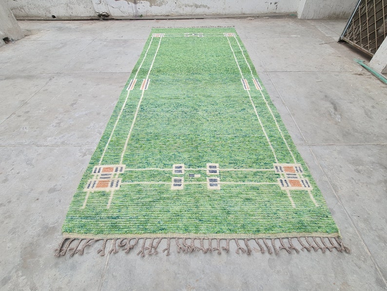 Puede incluir: Una alfombra rectangular verde con un dise&ntilde;o geom&eacute;trico. La alfombra presenta un campo verde claro con detalles en blanco, naranja y azul marino. La alfombra tiene flecos en los extremos cortos. La alfombra mide unos 152 cm de largo.