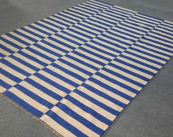 May include: A rectangular rug with a blue and off-white striped pattern. The stripes run horizontally and vertically, creating a checkered design. The rug is made of a woven material and is placed on a gray surface.