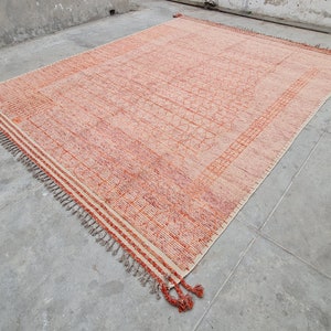 Puede incluir: Una alfombra rectangular con un patrón geométrico melocotón y naranja. La alfombra tiene un borde crema con rayas naranjas y borlas en los extremos. La alfombra está hecha de material tejido y se exhibe sobre un suelo de hormigón.