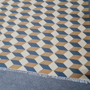 May include: Geometric patterned rug featuring a repeating 3D cube design in blue, tan, and off-white. The rug has a fringed edge and is suitable for various interior spaces.