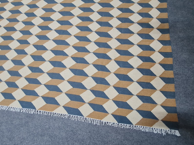 May include: Geometric patterned rug featuring a repeating 3D cube design in blue, tan, and off-white. The rug has a fringed edge and is suitable for various interior spaces.