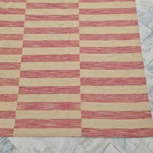 May include: A rectangular rug with a repeating pattern of horizontal stripes in alternating shades of beige and red. The rug is made of a woven material and has a simple, modern design. The rug is suitable for indoor use.