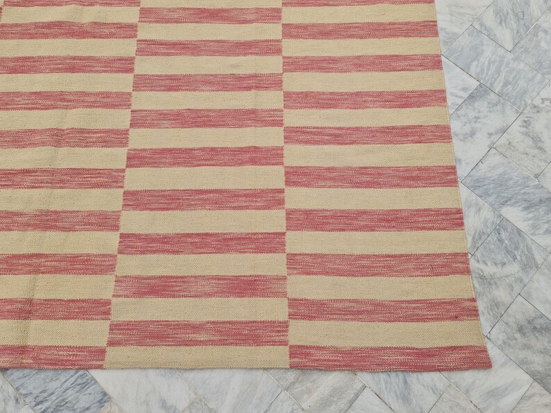 May include: A rectangular rug with a repeating pattern of horizontal stripes in alternating shades of beige and red. The rug is made of a woven material and has a simple, modern design. The rug is suitable for indoor use.