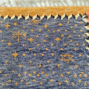 May include: A section of a blue rug with a star pattern in gold. The rug has a border of orange, black, and white geometric shapes. The top edge has a tan fringe.