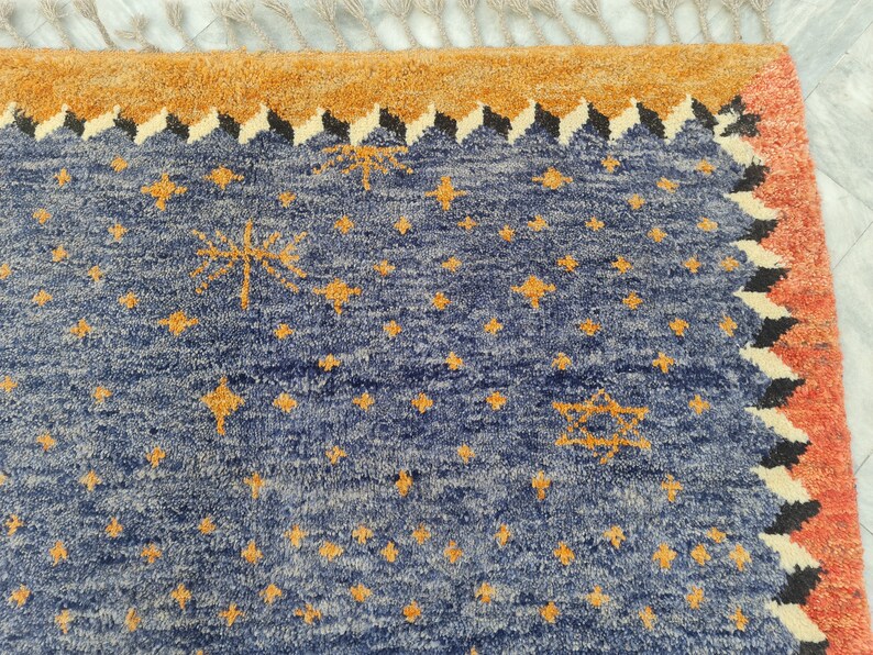 May include: A section of a blue rug with a star pattern in gold. The rug has a border of orange, black, and white geometric shapes. The top edge has a tan fringe.