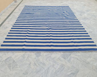 May include: A rectangular rug with horizontal stripes in alternating shades of blue and off-white. The rug has a fringed edge and is placed on a light-colored tiled surface. The rug's design is simple and modern, suitable for various interior styles.