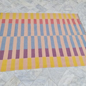 May include: A rectangular rug with a geometric pattern. The rug features vertical stripes in alternating colors of yellow, pink, blue, and purple. The rug has a fringed edge and is displayed on a marble floor.