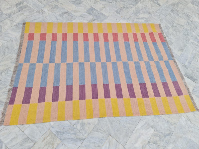 May include: A rectangular rug with a geometric pattern. The rug features vertical stripes in alternating colors of yellow, pink, blue, and purple. The rug has a fringed edge and is displayed on a marble floor.