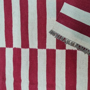 Puede incluir: Una alfombra a rayas rojas y blancas con una secci&oacute;n doblada a juego. La alfombra presenta rayas verticales, con un patr&oacute;n de tablero de ajedrez en la parte inferior. La secci&oacute;n doblada tiene un borde con flecos. La alfombra est&aacute; hecha de material tejido.