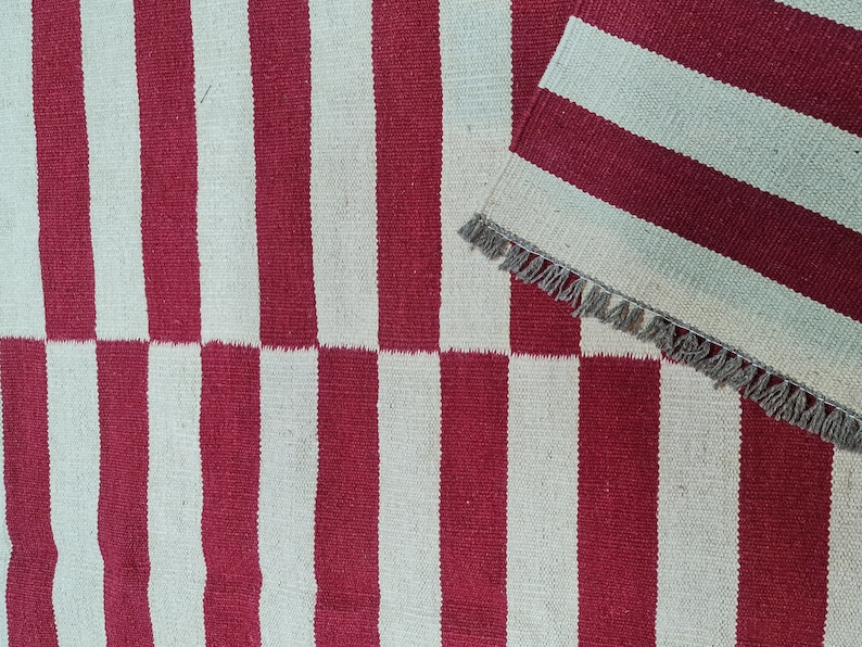 Puede incluir: Una alfombra a rayas rojas y blancas con una secci&oacute;n doblada a juego. La alfombra presenta rayas verticales, con un patr&oacute;n de tablero de ajedrez en la parte inferior. La secci&oacute;n doblada tiene un borde con flecos. La alfombra est&aacute; hecha de material tejido.