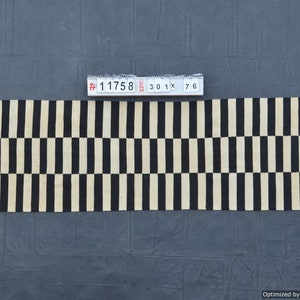 May include: A rectangular rug with a repeating pattern of black and beige vertical stripes. The rug is approximately 30 inches wide and 76 inches long. The rug is made of a woven material and is displayed on a dark gray surface.