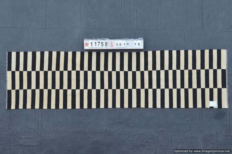 May include: A rectangular rug with a repeating pattern of black and beige vertical stripes. The rug is approximately 30 inches wide and 76 inches long. The rug is made of a woven material and is displayed on a dark gray surface.