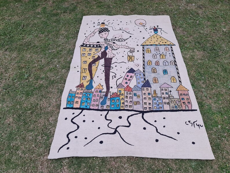 Op de afbeelding: Beige tapijt met een grillige illustratie van een persoon die door een stad loopt. Het kunstwerk toont kleurrijke gebouwen, een figuur met een hoed en een sterrenhemel. Het tapijt heeft een zwarte rand en de handtekening van de kunstenaar.