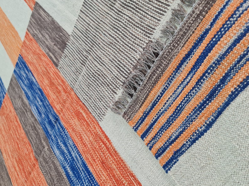 May include: A colorful woven rug with a geometric pattern. The rug features stripes in orange, blue, brown, and white. The rug has a fringed edge and is made of a textured material.