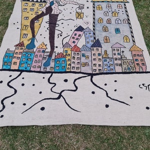 Op de afbeelding: Een handgeschilderd tapijt met een gestileerde stadsgezicht met kleurrijke gebouwen en een figuur met een lantaarn. Het kunstwerk bevat zwarte lijnen, stippen en de handtekening van de kunstenaar. Het tapijt ligt op groen gras.
