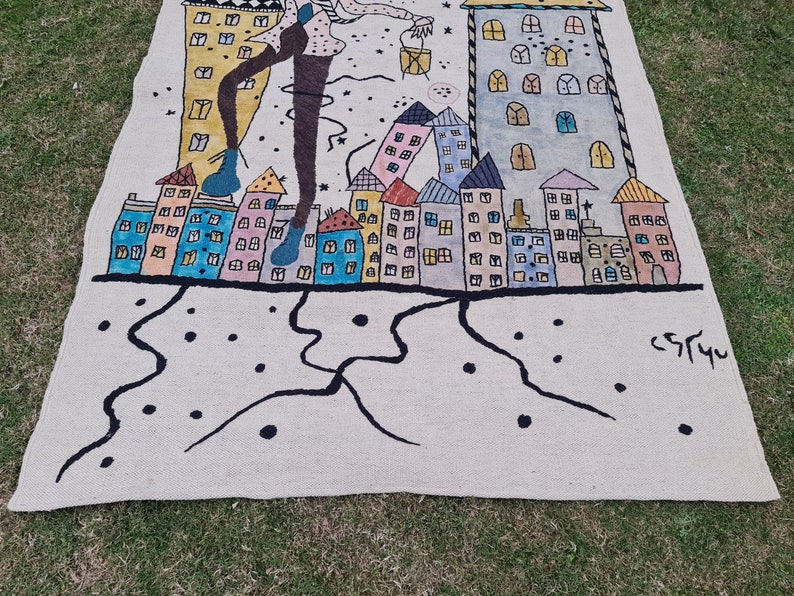 Op de afbeelding: Een handgeschilderd tapijt met een gestileerde stadsgezicht met kleurrijke gebouwen en een figuur met een lantaarn. Het kunstwerk bevat zwarte lijnen, stippen en de handtekening van de kunstenaar. Het tapijt ligt op groen gras.