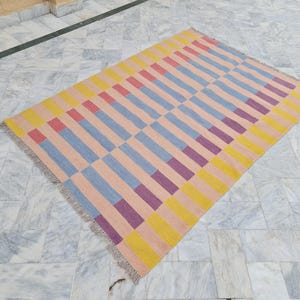 May include: Rectangular area rug with a geometric pattern. The rug features alternating rows of vertical stripes in peach, yellow, blue, red, and purple. The rug has a fringed edge and is placed on a marble floor.