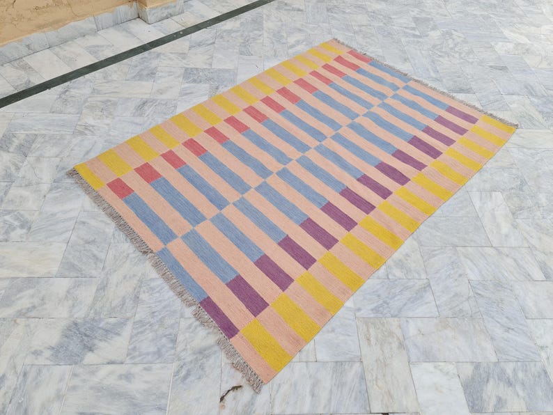 May include: Rectangular area rug with a geometric pattern. The rug features alternating rows of vertical stripes in peach, yellow, blue, red, and purple. The rug has a fringed edge and is placed on a marble floor.