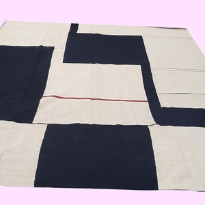 Op de afbeelding: Een groot, rechthoekig vloerkleed met een geometrisch ontwerp. Het vloerkleed heeft een patchworkpatroon van zwarte en gebroken witte rechthoekige vormen. Een dunne rode lijn accentueert het gebroken witte gedeelte. Het vloerkleed is gemaakt van een geweven materiaal.