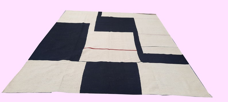 Op de afbeelding: Een groot, rechthoekig vloerkleed met een geometrisch ontwerp. Het vloerkleed heeft een patchworkpatroon van zwarte en gebroken witte rechthoekige vormen. Een dunne rode lijn accentueert het gebroken witte gedeelte. Het vloerkleed is gemaakt van een geweven materiaal.