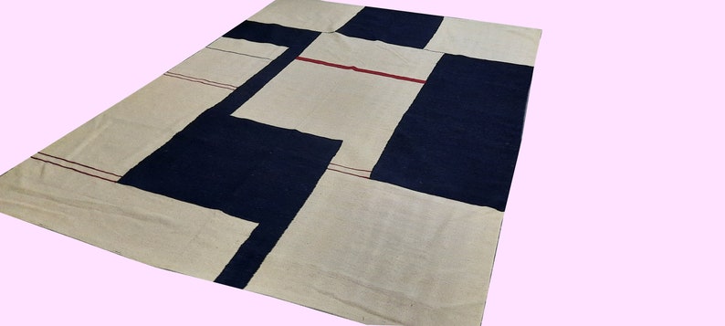 Op de afbeelding: Een groot rechthoekig tapijt met een geometrisch ontwerp. Het tapijt heeft cr&egrave;me, marineblauwe en rode accenten. Het ontwerp omvat grote kleurvlakken en dunne rode lijnen. Het tapijt is gemaakt van geweven materiaal.