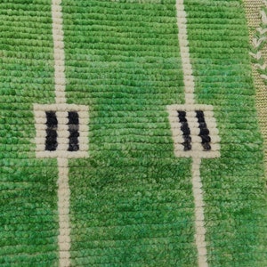 May include: Green rug with a football field design. The rug features a textured, grassy appearance with white lines and black and white end zone markings. The border has a decorative pattern in shades of green and beige.