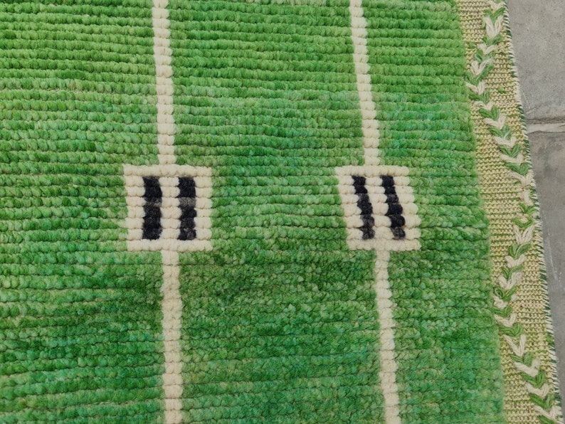 May include: Green rug with a football field design. The rug features a textured, grassy appearance with white lines and black and white end zone markings. The border has a decorative pattern in shades of green and beige.