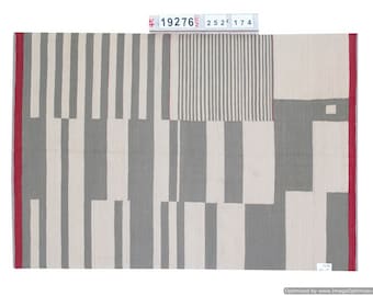 Puede incluir: Textil rectangular con un patrón geométrico. El diseño presenta rayas verticales en gris y blanco roto, intercaladas con bloques grises y blanco roto sólidos. Un borde rojo enmarca los bordes.