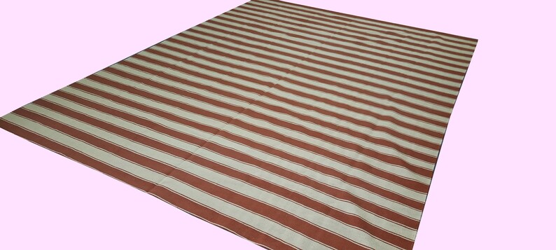 May include: A large, rectangular rug with a repeating pattern of wide, parallel stripes in alternating shades of brown and off-white. The rug is made of a woven material and is suitable for indoor or outdoor use.