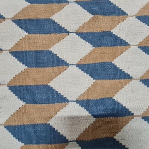 May include: A woven rug with a geometric pattern of diamonds in blue, tan, and white. The design creates a three-dimensional effect. The rug is made of a textured material, suitable for home decor.