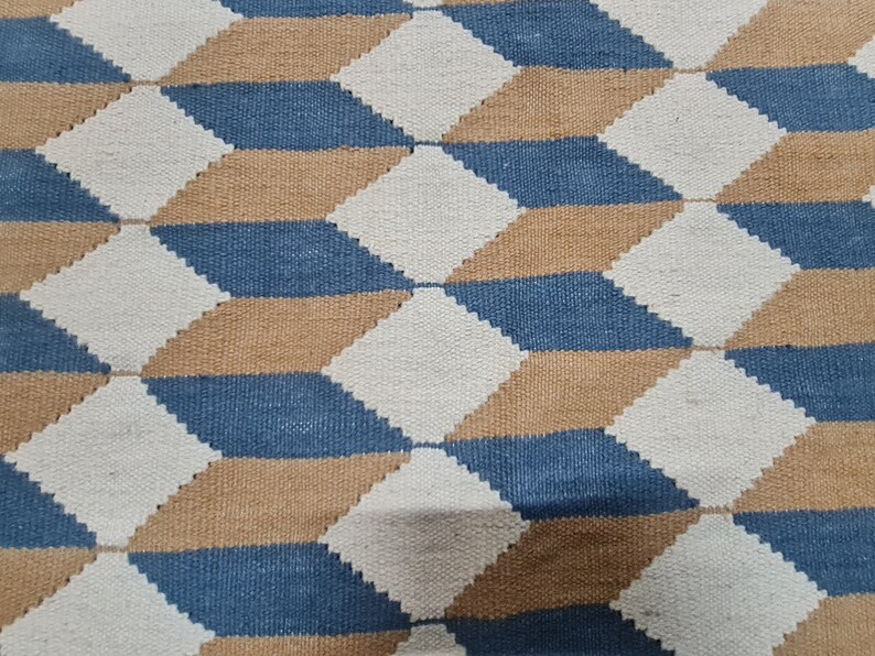 May include: A woven rug with a geometric pattern of diamonds in blue, tan, and white. The design creates a three-dimensional effect. The rug is made of a textured material, suitable for home decor.