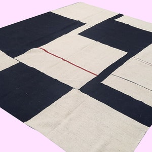 Op de afbeelding: Een groot, vierkant tapijt met een geometrisch patroon. Het tapijt heeft afwisselende blokken marineblauw en gebroken wit. Een dunne rode lijn accentueert de gebroken witte secties. Het tapijt is gemaakt van een geweven materiaal.