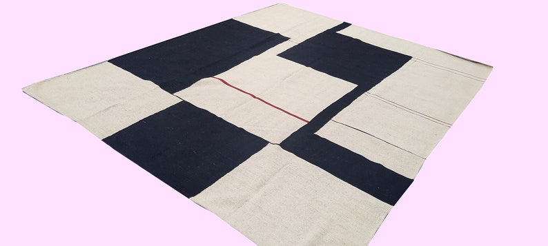 Op de afbeelding: Een groot, vierkant tapijt met een geometrisch patroon. Het tapijt heeft afwisselende blokken marineblauw en gebroken wit. Een dunne rode lijn accentueert de gebroken witte secties. Het tapijt is gemaakt van een geweven materiaal.