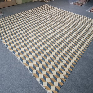 May include: Large geometric area rug featuring a repeating cube pattern in beige, blue, and brown. The rug has a woven texture and a fringed edge. The rug is rectangular and covers a large area.