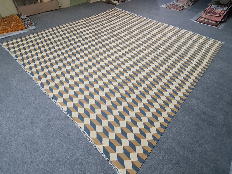 May include: Large geometric area rug featuring a repeating cube pattern in beige, blue, and brown. The rug has a woven texture and a fringed edge. The rug is rectangular and covers a large area.