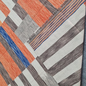 May include: Geometric patterned rug with a patchwork design. The rug features a combination of orange, blue, gray, and white stripes and blocks. The rug has a fringed edge and is made of woven material.
