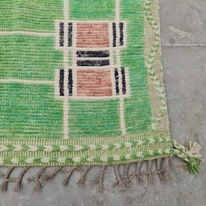 May include: A green and beige woven rug with a geometric pattern. The rug features a grid design with rectangular blocks in shades of green, beige, black, and pink. The rug has a fringe along the bottom edge.