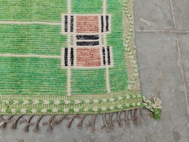May include: A green and beige woven rug with a geometric pattern. The rug features a grid design with rectangular blocks in shades of green, beige, black, and pink. The rug has a fringe along the bottom edge.