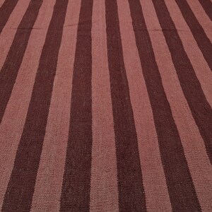 May include: A woven rug with a striped pattern. Alternating wide stripes of dark burgundy and muted rose create a bold, yet simple design. The rug's texture suggests a natural fiber construction, suitable for various interior styles.
