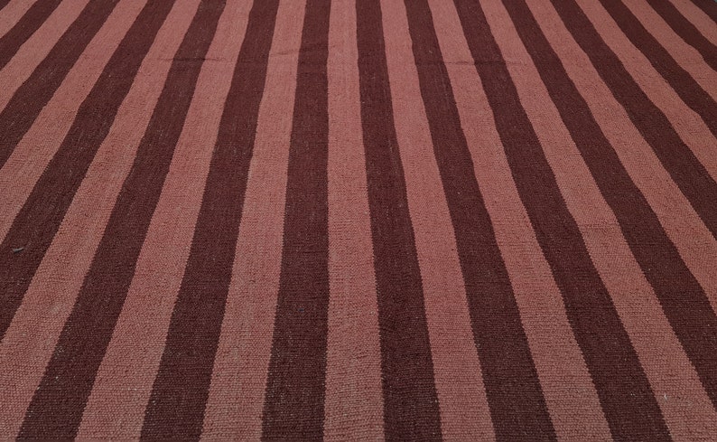 May include: A woven rug with a striped pattern. Alternating wide stripes of dark burgundy and muted rose create a bold, yet simple design. The rug's texture suggests a natural fiber construction, suitable for various interior styles.