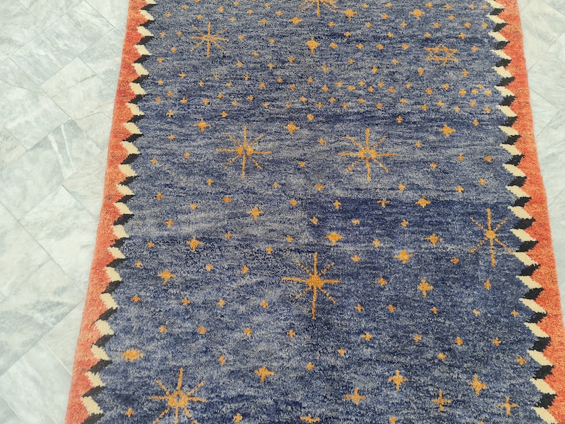 May include: A blue rug with a pattern of yellow starbursts and smaller stars. The rug has a red border with a zigzag design in black and white. The rug is made of a textured material.