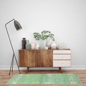Puede incluir: Un aparador de madera de estilo moderno de mediados de siglo con tres cajones y una alfombra verde con l&iacute;neas blancas. El aparador contiene varios jarrones, velas y vegetaci&oacute;n. Una l&aacute;mpara de pie negra est&aacute; a la izquierda.
