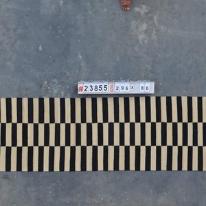 May include: A rectangular rug with a repeating pattern of black and beige vertical stripes. The rug measures 296 cm long and 80 cm wide. The rug is on a concrete surface.