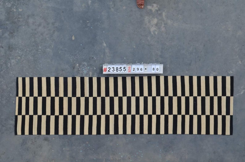 May include: A rectangular rug with a repeating pattern of black and beige vertical stripes. The rug measures 296 cm long and 80 cm wide. The rug is on a concrete surface.