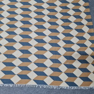 May include: Geometric patterned rug featuring a repeating cube design in blue, tan, and off-white. The rug has a fringed edge and a textured appearance, suitable for home decor.